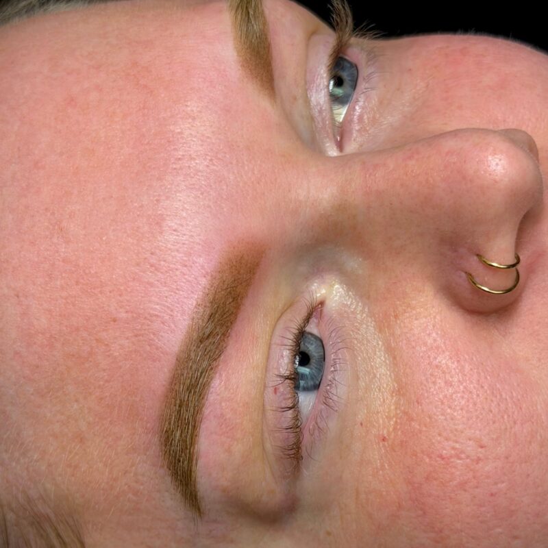 Healed permanent makeup brows by Lindsey on a client with blue eyes, photographed against a black backdrop.
